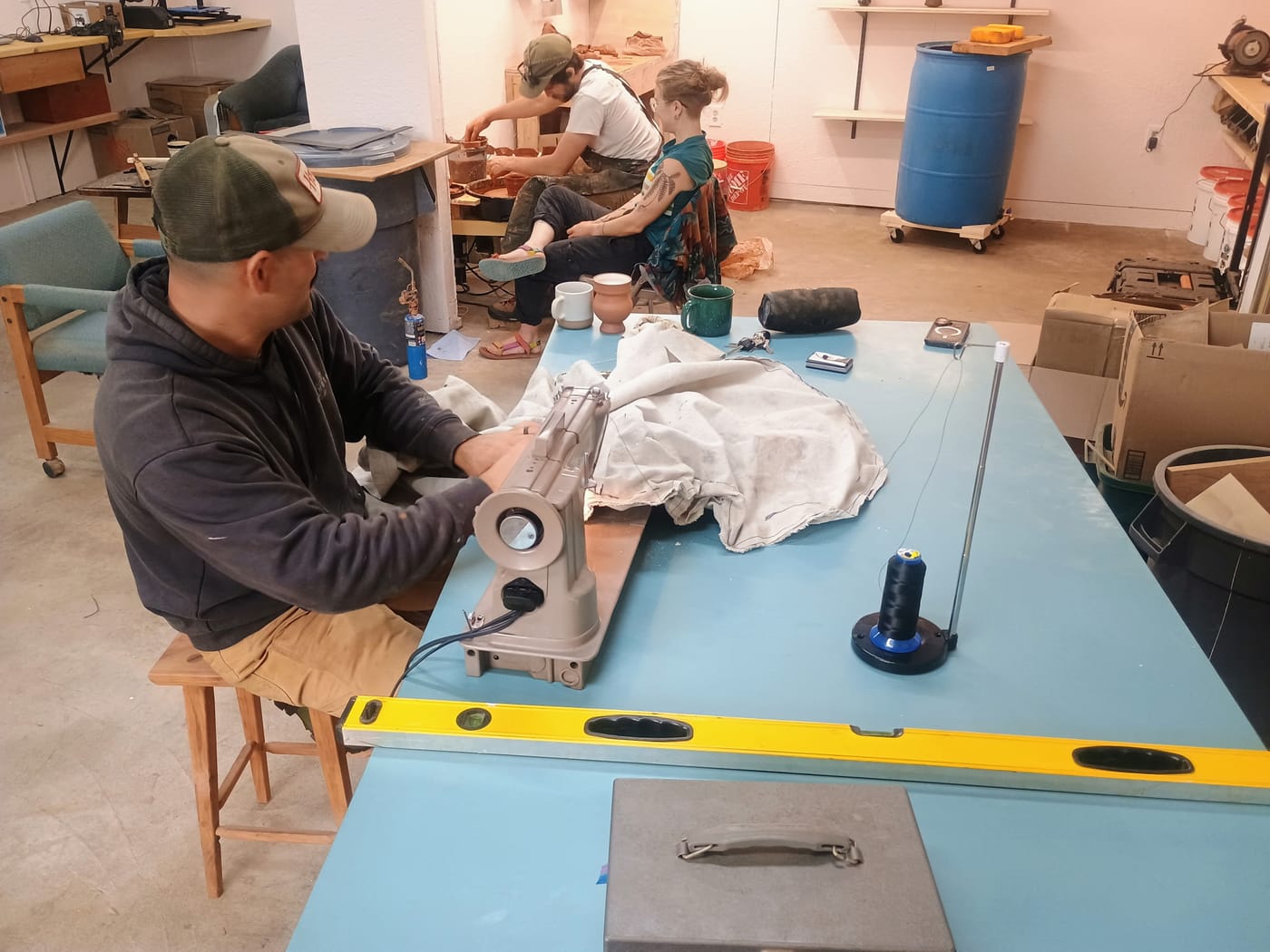 One man in the foreground working at a sewing machine, a man and a woman behind him are at a pottery wheel